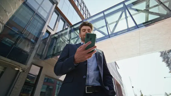 Confident entrepreneur looking cellphone screen on sunny street bottom view. Elegant businessman reading phone message standing urban area. Handsome company owner browsing smartphone app outdoors.