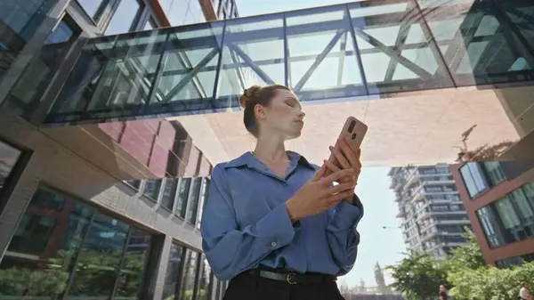 Businesswoman checking smartphone email on sunny urban street bottom view. Confident woman boss looking cellphone standing under modern office construction. Elegant lady scrolling mobile phone screen.