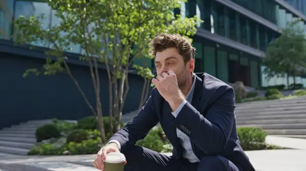Relaxed employee eating sandwich sitting street stairs with beverage takeaway. Formal suit manager relaxing at lunch break outside office. Handsome man freelancer holding coffee cup outdoors zoom out.