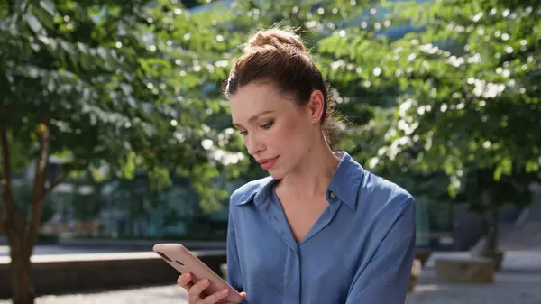 Business lady looking smartphone screen relaxing in sunny town park close up. Serious female freelancer browsing cellphone app sitting alley with green trees. Confident businesswoman reading message.