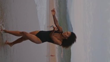Afrika kökenli Amerikalı kadın bulutlu plajda dans ediyor. Mayo giymiş dikey bir melek. İnce kıvırcık kız ıslak kumda yürürken yumuşak eller hareketi yapıyor. Seksi esmer kasvetli gökyüzünde gösteri yapıyor.