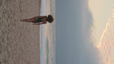 Rahatlamış genç bir kadın akşam manzarasında okyanus dalgalarının yanında kumsalda yürümenin tadını çıkarıyor. Siyah mayo giymiş bilinmeyen kıvırcık esmer deniz videosuna gidiyor. Sıska kız bulutlu gökyüzünde dinleniyor