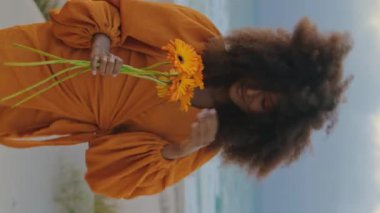 Afro-amerikan neşeli kız, rüzgarlı kasvetli sahilde dikey turuncu buketle poz veriyor. Kıvırcık saçlı çekici bir kadın. Gülümseyen bir kamera. Kumsalda çiçeklerle duruyor..