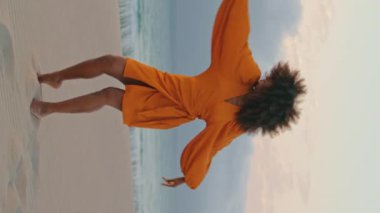 Çağdaş dansçı kız, kum kıyısı yaz akşamında el sallıyor. Gülümseyen esnek Afrikalı Amerikalı kadın turuncu elbise giyiyor kasvetli plajda modern dans tarzı..