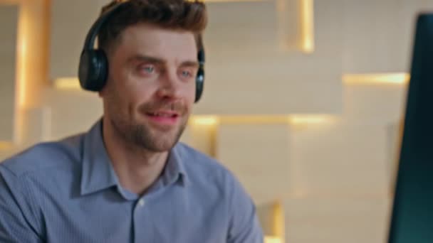 クリエイティブオフィスでキーボードを入力する男が閉じます デスクトップコンピュータで音楽を聴くワイヤレスヘッドフォンのハッピービジネスマン イヤホンの笑顔の会社のマネージャーは職場でサウンドを楽しむ — ストック動画