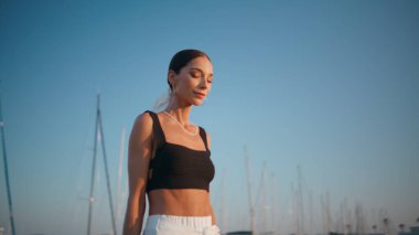 Gün batımı rıhtımında yürüyen muhteşem kadın kameraya güvenle bakıyor. Şık, lüks bir bayan yatların yanında akşam seti etrafında geziniyor. Güzel yaz günbatımında limana giden zarif genç kız..