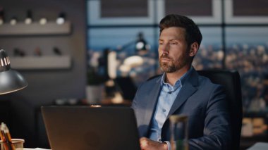 Kapalı mekanda kahve içen çok çalışan adam. Ciddi bir ofis çalışanı bilgisayar monitörünün çalışma alanına bakıyor. Sakallı, odaklanmış bir adam, akşam panoramik pencerelerde çay fincanının keyfini çıkarıyor.