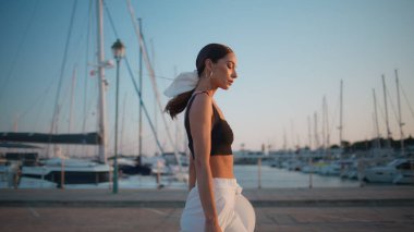 Beyaz lüks yatların yanında günbatımı seti gezen çekici bir model. Şık, kendine güvenen bir kadın, güneşli bir rıhtımda son moda kıyafetleriyle yürüyor. Muhteşem kız asfalt sahil yoluna çıkıyor yaz akşamı.