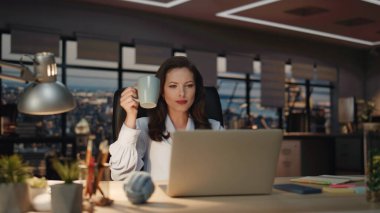 Kapuçino fincanının keyfini çıkaran tatmin olmuş bir bayan akşam geç saatlerde iş yerinde yakın çekim yapıyor. Rahat bir iş kadını, laptopunda kahve içiyor ve dinleniyor. Patron işkolik panoramik gece görüşüne ara verdi 