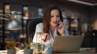 Geniş pencereli karanlık ofiste pozitif yönetici cep telefonuyla konuşuyor. Duygusal iş kadını akıllı telefonu arıyor ve akşam dolabına gülüyor. Gülümseyen bayan patron cep telefonu şakalarına tepki veriyor.