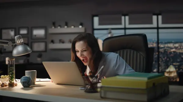 Woman winner looking laptop celebrating success in evening office. Happy boss screaming loudly enjoying business achievement. Emotional lady gesturing hands exciting by good news in dark cabinet