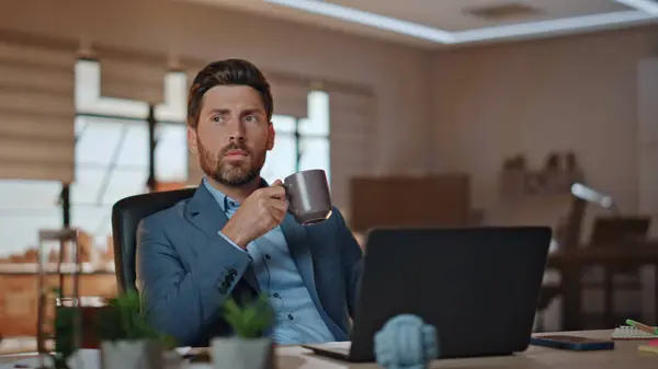 Relaxed manager holding coffee cup at light workplace closeup. Serious bearded man taking break working at modern office interior. Calm rich businessman looking laptop computer sitting at desk alone