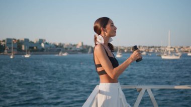 Dürbünlü kadın yaz gezisinde deniz güzelini izliyor. Güneşli rıhtımda duran mutlu çekici kız turist güzel deniz manzarasının tadını çıkarıyor. Moda tebessümlü esmer bayan kamera kıyıda poz veriyor..