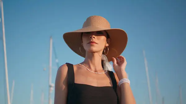 Elegant hat model posing in front yachts masts at embankment close up. Portrait of luxurious posh woman standing at beautiful blue sky. Gorgeous confident girl looking camera enjoying summer vacation.