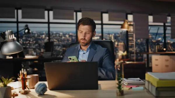 Disappointed manager reading message on smartphone sitting at night luxury office with panoramic window closeup. Bearded businessman feeling stress receiving bad news on phone. Ceo worried work