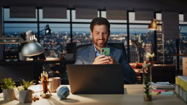 Happy boss relaxing with cellphone at late office closeup. Positive businessman watching funny videos smiling toothy at break in dark workplace. Cheerful manager chatting friend at mobile phone alone