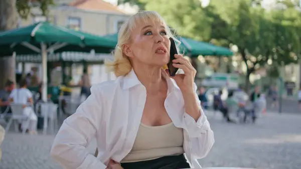 Old lady speaking smartphone at restaurant terrace close up. Serious senior businesswoman have work conversation by cellphone outdoors. Elegant aged woman calling phone sitting table street cafeteria.