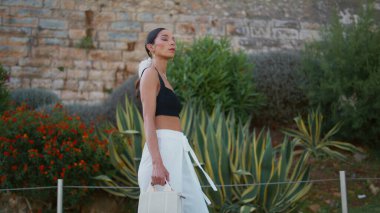 Yeşillik görünümlü kamerasıyla parkta yürüyen muhteşem bir kadın. Zarif, ciddi bir kadın, tropikal caddelerde moda kıyafeti giyerek dolaşıyor. Genç zengin esmer, yeşil sokakta randevuya çıkıyor. Boş zaman konsepti.