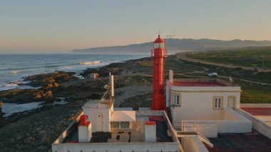 Manzaralı deniz feneri drone görüntüsü. Güzel kırmızı kule feneri kayalık deniz kıyısında yaz günü duruyor. Resimli okyanus dalgaları kıyı kayalıklarına çok yavaş bir şekilde çarpıyor. Sakin deniz manzarası.