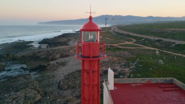 Drone manzaralı deniz feneri akşam sahilinde parlıyor. Okyanus kıyısında alacakaranlıkta kırmızı fener yanıp sönüyor. Yüksek kule gemi navigasyonuna ışık sinyali veriyor. Resimli deniz manzarası.