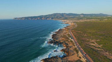Asfalt yol güneşli yaz günü deniz manzaralı. Güzel okyanus dalgaları çok yavaş çekimde engebeli kıyı şeridine çarpıyor. Düz yolda giden çeşitli arabalar. Tatil gezisi konsepti.