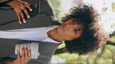 Düşünceli bir iş kadını elinde kağıt kahve fincanıyla şehir parkında akıllı bir telefon gibi duruyor. Kendine güvenen Afro-Amerikalı şık takım elbiseli kız telefonu dikey odaklı düşüncelere kaydırıyor..