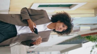 Akıllı telefon kullanan bir kadın şık bir takım elbiseyle caddede dikey bir şekilde yürüyor. Güzel Afro-Amerikalı kız telefon ekranında mesaj yazıyor. Mutlu kıvırcık iş kadını cep telefonuyla çalışıyor..