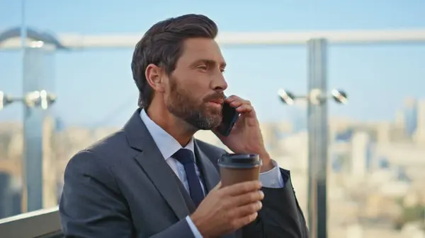 Bearded man sipping coffee at blurred cityscape closeup. Smiling manager calling mobile phone discussing project with partner on terrace. Relaxed boss executive drinking cup enjoying cell conversation
