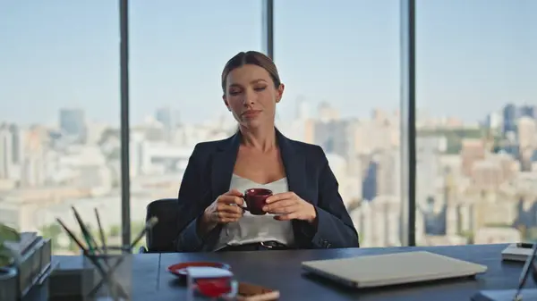 Company executive holding cup resting at office desk. Worried woman thinking problem waiting meeting at panoramic city view. Beautiful thoughtful businesswoman taking break relaxing at workplace.