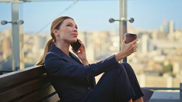 Relaxed businesswoman calling smartphone on glass terrace. Smiling manager rest on office balcony barefoot holding coffee at city view. Successful worker taking break having mobile phone conversation.