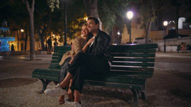 Parktaki bankta oturmuş, akşam randevusunda kucaklaşan sevimli bir çift. Mutlu eşler gece kenti birlikte dinleniyor. Gülümseyen yakışıklı adam dışarıda neşeli kadını kucaklıyor. Kentsel aşk ilişkisi konsepti.