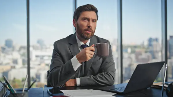 Calm businessman sipping coffee thinking solution at desk. Inspired manager typing laptop searching online at panoramic window. Thoughtful bearded man finishing break working computer in modern office