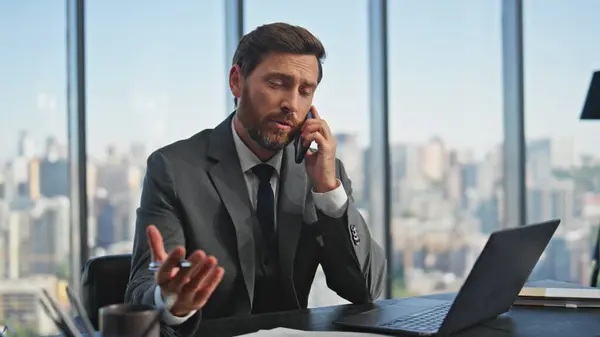 Business consultant talk smartphone in contemporary office. Confident ceo in suit working discussing financial project with partner on call at cityscape. Successful boss sitting desk speaking cell.