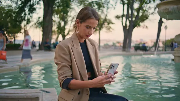 Joyful woman taking selfie video in city park. Beautiful girl call cell sending virtual kiss at water fountain. Happy stylish businesswoman using mobile phone recording social media message on street