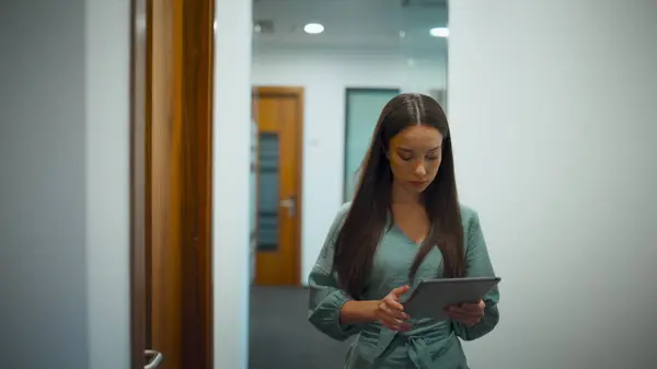 Serious employee working on go in office. Professional woman ceo using tablet computer browsing internet in hallway. Focused project manager walking glass corridor. Corporate busy lifestyle concept