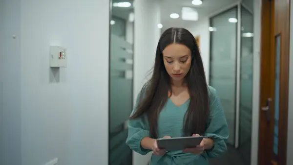 Woman ceo browsing tablet computer in office. Busy manager walking through corridor in modern workplace. Business person reading corporate data in glass wall hallway. Professional diverse concept