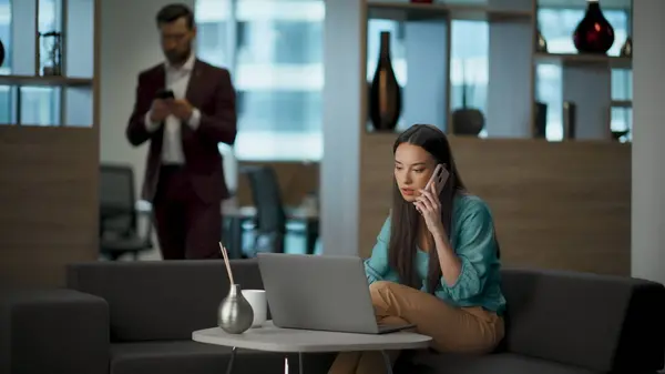 Focused team lead working laptop computer in office interior. Confident worker answering on phone call. Woman talking smartphone at remote workplace. Business person calling at luxury place
