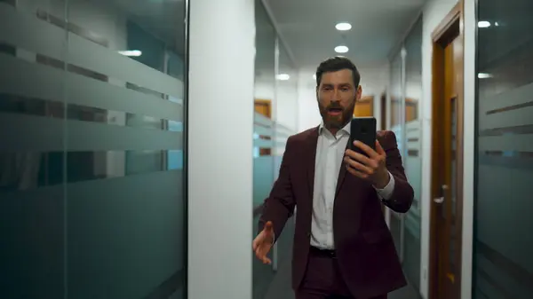Angry chief arguing video call conversation in business center. Nervous businessman holding smartphone in glass walls corridor. Closeup emotional boss walking chatting mobile phone in office hallway