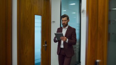 Yönetici ofisteki tablet bilgisayarı arıyor. Modern işyerindeki koridorda yürüyen sakallı bir yönetici. İş adamı cam koridorda şirket verilerine bakıyor. Profesyonel konsept 