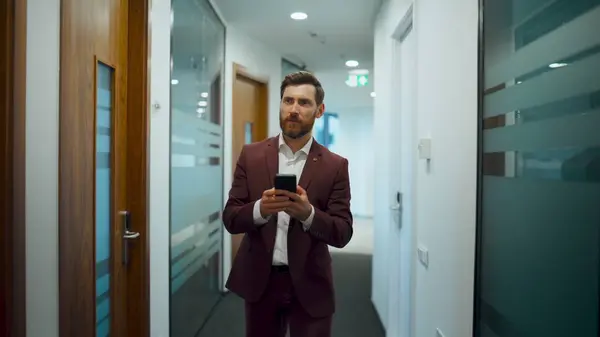 Successful businessman walking with phone at corridor. Confident bearded man using smartphone crossing through glass wall place. Serious professional using mobile inside. Career people concept