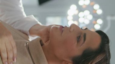 Çekici orta yaşlı kadın dikey olarak aynaya bakarak tatmin olmuş yüz tazelemesi yapıyor. Gülümseyen kozmetik uzmanı yaşlanmayı önleyici terapi uyguluyor. Cilt kaldırma kırışıklıkları düzeltmesi