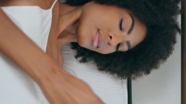 Yatakta özçekim yapan muhteşem bir manken. Afroamerikan rahat bayan cep telefonuyla fotoğraf çekiyor ve yastık bırakıyor. Seksi kız cep telefonu kamerası için saçlarını düzleştiriyor. Dikey görünüm sergileyen kaygısız kadın