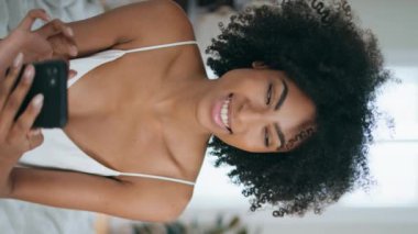 Gülümseyen kadın yatak odasında akıllı telefondan mesaj atıyor. Kıvırcık saçlı, gülen kadın cep telefonuyla mesaj atıyor. Afro-Amerikalı kız, yatağında oturmuş kamera görüntüsü çiziyor.. 