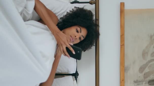 スマートフォン高級寝室を読み取る優しいモデル 美しいカーリー女性は 垂直方向に携帯電話を保持する白いインテリアで休憩します 自宅で電話をスクロールする魅力的なアフリカの少女 — ストック動画