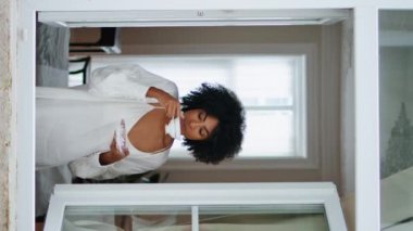 Rahat kadın, pencere çerçevesinde espresso fincanının keyfini çıkarıyor. Afrikalı Amerikalı kaygısız kadın yalnız başına dikey videonun dışına bakıyor. Güzel kıvırcık kız oturma odasında kahve fincanıyla dinleniyor. Günlük rutin 