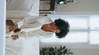 Sabah beyaz yatak odasında çorap giyen sakin bir kız. Kaygısız kadın model, yeni bir güne başlamak için yatağını dikey ve rahat bir şekilde hazırlıyor. Afrikalı Amerikalı romantik kadın günlük rutin işler yapıyor.. 