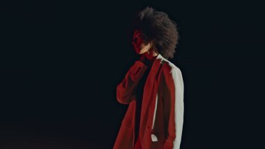 Model, sanat makyajlı, hafif görünümlü bir kameraya dönüşüyor. Siyah arka planda poz veren şenlikli kulüp kızı. Göz kamaştırıcı rüya gibi Afro-Amerikan gözlerini kapayarak saçlarına dokunuyor. Beyaz ceketli şehvetli kadın.