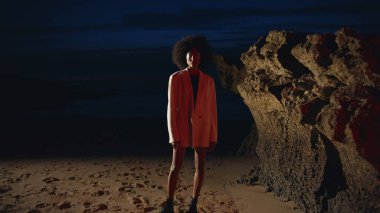 Geceleri ceketle Cliff Shore 'da modellik yapan seksi kız. Projektör ışığında kumlu kumsalda poz veren zayıf, çekici bir kadın. Kaygısız moda Afro-Amerikan dansı akşamın tadını çıkarıyor. Açık havada şehvetli bir kadın
