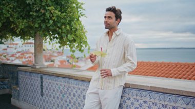 Bulutlu bir bakış açısında elinde çiçekle gülümseyen adam yakınlaş. Yakından mutlu, sakallı, deniz manzaralı yaz tatili manzarası seyreden adam. İspanyol esmer, kız arkadaşını bekliyor, dinleniyor.