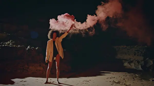 Fashion woman waving bomb on night beach. Protest girl holding smoke grenade walking sandy shore in light. Slim african american posing camera under colorful steam. Party model celebrating outdoors.